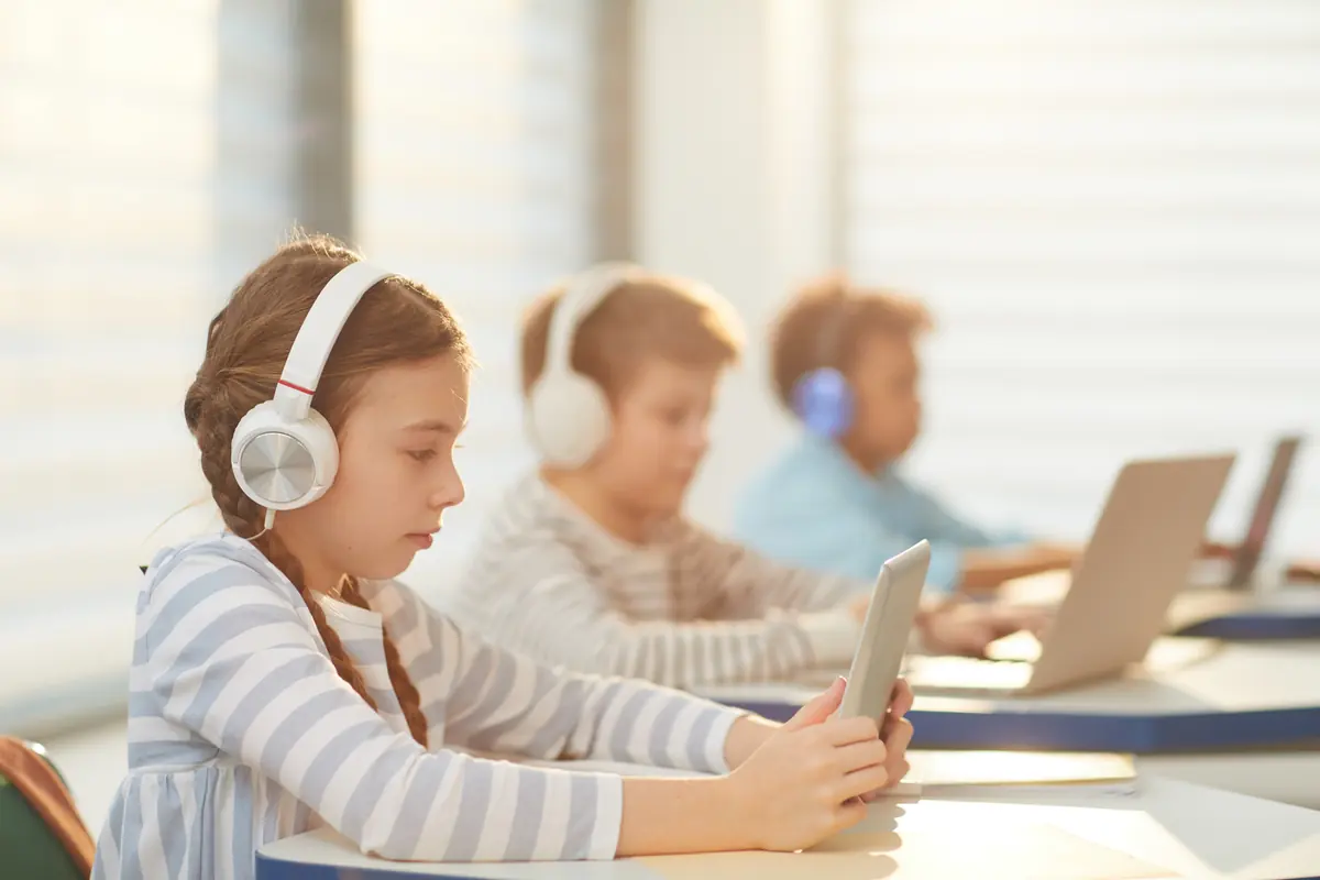 Students at classroom desks working on laptops and tablets with headphones